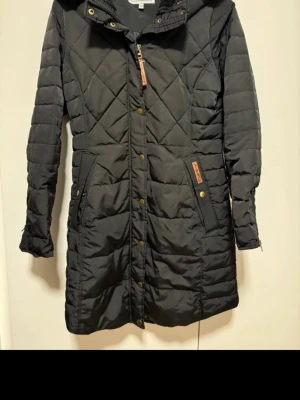 Svart quiltad dunjacka från Janbell - Lång svart dunjacka från Janbell med quiltat mönster, huva och mysig fuskpäls på insidan av kragen. Jackan har dragkedja, knappar i guldton och praktiska fickor med snygga detaljer. Perfekt för kalla dagar och riktigt skön passform.