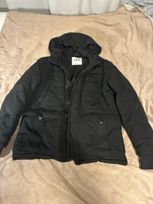 Svart parkajacka från Zara med huva - Säljer en svart parkajacka från Zara i storlek S. Jackan har huva, stora fickor med knappar och dragkedja framtill. Perfekt för kyligare dagar och har en klassisk, enkel design som funkar till det mesta. En knapp fattas längst ner, men jackan funkar fortfarande perfekt.