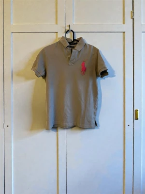 Vintage Beige Polo Ralph Lauren piké tröja - Säljer en grå kortärmad pikétröja från Polo Ralph Lauren i custom fit. Stor rosa logga på bröstet och en rosa siffra 3 på ärmen. Klassisk pikékrage och knappar framtill. Skön bomullskvalitet och snygg passform.