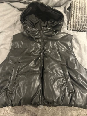 Svart dunväst med huva i skinnimitation - Säljer en svart dunväst med huva i skinnimitation. Västen har dragkedja framtill och två fickor på sidorna. Perfekt för lager på lager och ger en cool vibe till din outfit. Passar dig som gillar streetstyle och vill ha något både snyggt och praktiskt.