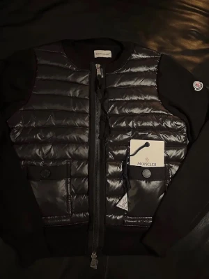 Moncler cardigan  - Säljer en svart pufferjacka från Moncler med quiltad front och två fickor med knappstängning. Jackan har dragkedja framtill och Moncler-logga på ärmen. Materialmix med blank polyester framtill och stickade ärmar och rygg. Perfekt för dig som vill ha en stilren och exklusiv look. Kommer med prislapp