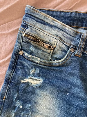 Blå distressed jeans Supply & Demand - Säljer ett par blå jeans från Supply & Demand med slitna detaljer och dragkedja vid fickan. Jeansen har en tvättad look, raka ben och klassisk femficksdesign. Materialet är jeans och passformen är normal. Perfekta för dig som gillar en avslappnad streetstil.