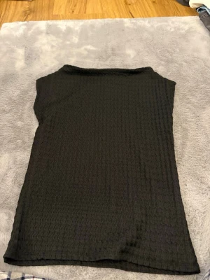 Svart ribbad topp XS - Säljer en svart ribbad topp i storlek XS. Toppen har ett vågigt ribbat mönster och är ärmlös med en enkel, rak passform. Perfekt att styla med jeans eller kjol för en clean look.