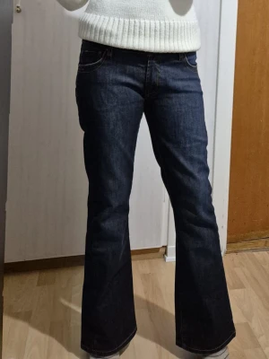 Mörkblå bootcut jeans Wrangler - Säljer ett par mörkblå bootcut jeans med klassiska kontrastsömmar och snygga bakfickor med gul brodyr. Jeansen har normal passform och är tillverkade i slitstarkt denimtyg. Perfekta för dig som gillar vintage kläder och vill ha ett par riktigt schyssta jeans. Är i utmärkt skick.