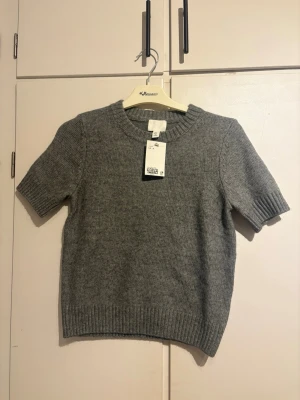 Grå stickad kortärmad tröja H&M - Säljer en enkel och stilren grå stickad tröja från H&M i storlek S. Tröjan har korta ärmar, rund halsringning och ribbade muddar vid ärmslut och nederkant. Perfekt att matcha med jeans eller kjol för en clean look.