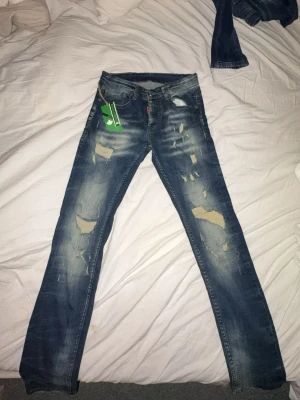 Dsquared2 blå slitna jeans, stl 30 - Säljer ett par blå jeans från Dsquared2 i storlek 30. Jeansen har en snygg sliten look med flera slitningar och lagningar framtill, klassisk femficksmodell och raka ben. Tillverkade i 100% bomull för skön känsla och cool stil.