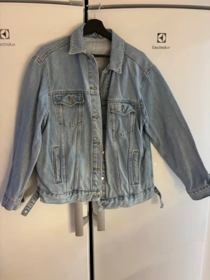 Ljusblå oversized jeansjacka - Säljer en klassisk ljusblå jeansjacka i oversized modell. Jackan har två bröstfickor med lock och knappar, samt sidofickor. Knappstängning framtill och justerbara spännen i midjan. Perfekt att slänga över en hoodie eller t-shirt.