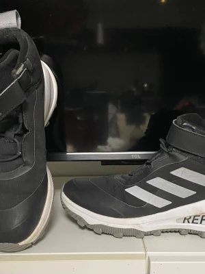 Svarta adidas sneakers strl 38 - Säljer ett par svarta adidas sneakers med vita detaljer och grå sula. Skorna har kardborreband upptill, klassiska tre ränder på sidan och grovt mönstrad yttersula för extra grepp. Tillverkade i syntetmaterial och textil, perfekta för utomhusaktiviteter.