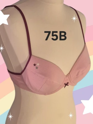 Rosa bh med vinröda detaljer 75B - Söt rosa bh med tunna vinröda axelband och kant. Dekorerad med en liten rosett framtill och små stjärnor på ena kupan. Justerbara axelband och klassisk hyskknäppning bak. Perfekt för dig som gillar en feminin och enkel stil. Uttagbar kuddar.