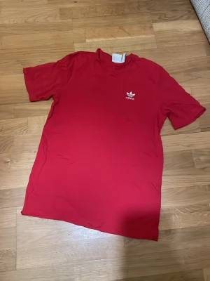 Röd Adidas t-shirt med logga - Säljer en röd t-shirt från Adidas med den klassiska treklöver-loggan tryckt i vitt på bröstet. T-shirten har rund hals och korta ärmar. Tillverkad i mjuk bomull som är skön att bära hela dagen storlek L passar som xs-S i herr.