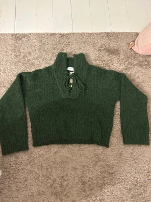 Mörkgrön stickad tröja från Vila - Mysig mörkgrön stickad tröja från Vila med hög krage och snörning framtill. Tröjan har ribbade muddar och är lite croppad i modellen. Perfekt för kyliga dagar och passar snyggt till jeans.