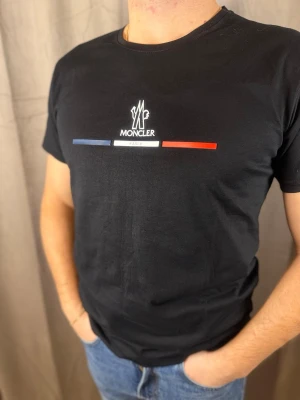 Svart Moncler t-shirt med tryck - Svart t-shirt från Moncler med klassiskt tryck på bröstet i blått, vitt och rött samt texten 'Paris'. T-shirten har rund hals och korta ärmar. Tillverkad i mjuk bomull som känns skön mot huden. Perfekt för dig som gillar stilrena och exklusiva plagg.