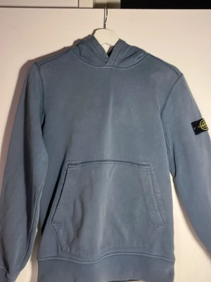 Blå Stone island hoodie storlek xs - Säljer min ljusblåa stone island hoodie med en unik design på ryggen. Hoodien har inga defekter men har ett använt skick då den används länge. 