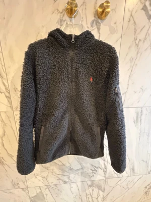 Svart teddyjacka från Polo Ralph Lauren - Mysig svart teddyjacka från Polo Ralph Lauren med huva och dragkedja framtill. Jackan har två fickor, en liten ficka med dragkedja på ärmen och den klassiska röda Polo-loggan broderad på bröstet. Perfekt för kalla dagar och riktigt skön att bära.