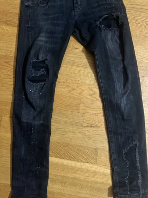 Svarta skinny jeans med slitningar - Säljer ett par svarta skinny jeans från Zara med slitna detaljer och hål på knäna och låren. Jeansen har en tvättad look och små vita stänk på både fram- och baksida. Klassisk femficksmodell och stretchigt material för skön passform. Hållen e lite öppnade men man kan sy dem o de e lite varma deme bra o fina som streetwear