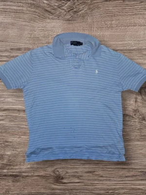 Randig piké från Polo Ralph Lauren - Ljusgrå pikétröja från Polo Ralph Lauren med tunna vita ränder och klassisk krage. Ikoniska broderade loggan på bröstet. Tillverkad i mjuk bomull och har korta ärmar. Perfekt för en avslappnad och stilren look.