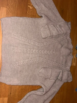 Beige stickad tröja med volang Vero Moda - Mysig beige stickad tröja från Vero Moda med flätstickat mönster framtill och volangdetaljer på axlarna. Tröjan har lång ärm och ribbade muddar vid hals, ärmslut och nederkant. Perfekt för kyliga dagar och enkel att styla. Har använt en gång 