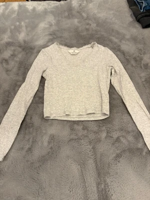 Ljusgrå ribbad topp från H&M - Ljusgrå långärmad topp från H&M med v-ringning och spetsdetalj runt halsen. Toppen är ribbad och har en croppad passform som ger en modern look. Perfekt att matcha med jeans eller kjol.
