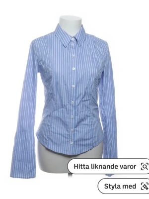 Blåvit randig skjorta med knappar - Säljer en klassisk blå skjorta med vita vertikala ränder. Skjortan har lång ärm, krage och knäppning framtill. Perfekt för dig som gillar stilrena och tidlösa plagg. Tillverkad i bomull för en skön känsla.