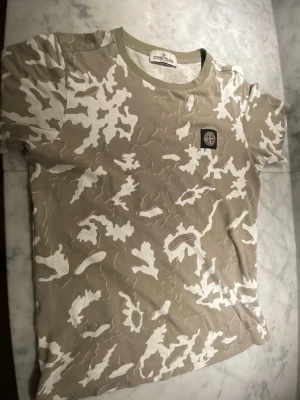 Stone Island Junior camo t-shirt - Snygg t-shirt från Stone Island Junior med beige och vitt camouflage-mönster. Klassisk rund halsringning med ribbad kant och ikonisk Stone Island-patch på bröstet. Perfekt för dig som vill sticka ut med streetwear-vibe.