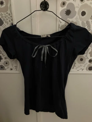 Mörkblå topp med rutigt knytband - Säljer en mörkblå topp med korta ärmar och har ett sött rutigt knytband, man knuter rosetten själv. Den är från 2000talet men i riktigt bra skick!