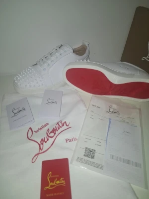 Vita Louboutin  - Säljer ett par vita sneakers från Christian Louboutin med ikoniska röda sulor och coola nitar över tån. Skorna har snörning och är tillverkade i skinn för en lyxig känsla. Perfekta för dig som vill sticka ut med exklusiv design och edgy detaljer. Pris går att diskuteras! Fraktar samma dag som köpet!