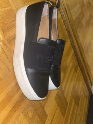 Svarta slip-on sneakers Calvin Klein - Snygga slip-on sneakers från Calvin Klein i svart skinn med bred resår över foten där loggan är präglad. Skorna har vit, tjock platåsula och rund tå. Perfekta för en clean och modern look.