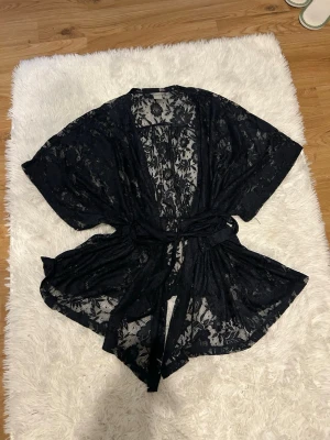 Svart spetsväst från Lindex - Säljer en svart, transparent spetsväst från Lindex i storlek L. Västen har ett vackert blommigt spetsmönster, korta ärmar och knytskärp i midjan för en snygg siluett. Perfekt för att lyfta vilken outfit som helst.