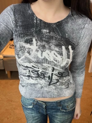 Grå croppad långärmad topp - Säljer en gråmelerad croppad långärmad topp med coolt abstrakt tryck och vita detaljer framtill. Toppen har rund halsringning och är dekorerad med små silvriga nitar som ger extra edge. Perfekt till ett par jeans för en avslappnad men trendig look.