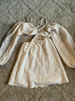 Beige och vit kortklänning H&M XS - Supersöt kortklänning från H&M i beige och vitt med diskret rutigt mönster. Klänningen har långa puffärmar, cut-out detalj framtill och smala axelband. Tillverkad i ett lätt och luftigt material, perfekt för varma dagar.