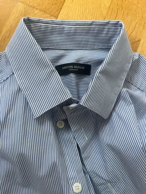 Randig skjorta från Bruuns Bazaar - Säljer en klassisk skjorta från Bruuns Bazaar i storlek L. Skjortan är blå- och vitrandig med långa ärmar, knappar framtill och bred krage. Perfekt för dig som gillar stilrena och tidlösa plagg. Materialet känns lätt och andas bra.