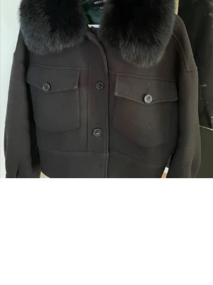 Svart kappa  från Meotine  - Säljer en svart kappa från Meotine med stor fluffig krage i päls och stora bröstfickor med lock och knappar. Västen har en rak, croppad passform och stängs med knappar framtill. Perfekt för dig som vill ha en stilren och trendig look.