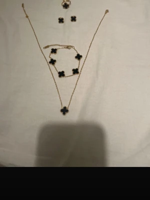 Svart och guld fyrklöver smyckesset - Säljer ett snyggt smyckesset med halsband, armband, ring och örhängen i svart och guld. Alla delar har fyrklöverformade hängen med guldkant. Haösband, armband, örhängen och ring. Perfekt för dig som gillar stilrena accessoarer med en touch av lyx.