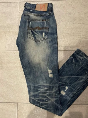 Nudie Jeans blå slitna jeans W28 L32 - Säljer ett par blå jeans från Nudie Jeans med slitningar och distressed detaljer. Klassisk femficksmodell med broderad bakficka och läderpatch i midjan. Jeansen har en rak passform och är tillverkade i slitstarkt denimtyg.