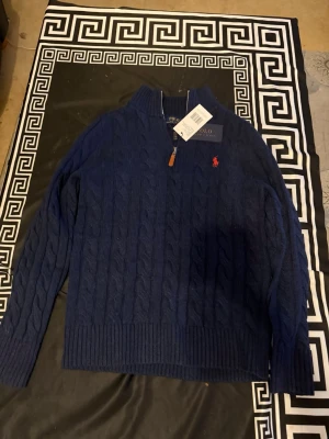 Marinblå stickad tröja Polo Ralph Lauren - Säljer en marinblå kabelstickad tröja från Polo Ralph Lauren med half zip och ribbade muddar. Tröjan har lång ärm och den klassiska röda loggan broderad på bröstet. Perfekt för dig som gillar stilren och tidlös design.
