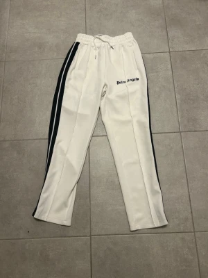 Vita Palm Angels mjukisbyxor med svarta ränder - Säljer ett par vita mjukisbyxor från Palm Angels med svarta ränder längs sidorna och logga på vänster lår. Byxorna har resårmidja med snörning och raka ben. Tillverkade i ett mjukt syntetmaterial som ger en sportig vibe.