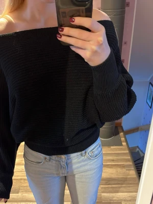 Svart offshoulder stickad tröja - Säljer en svart stickad tröja med offshoulder-design och långa ärmar. Tröjan har en loose passform och bred ribbad mudd vid ärmslut och nederkant. Perfekt för dig som gillar en avslappnad men ändå trendig stil.