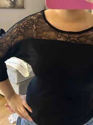 Svart topp med spetsdetaljer - Säljer en svart topp med genombruten spets över axlar och ärmar. Toppen har trekvartsärm och en rund halsringning. Spetsen har ett blommigt mönster som ger en elegant touch. Passformen är normal och materialet känns mjukt och stretchigt.