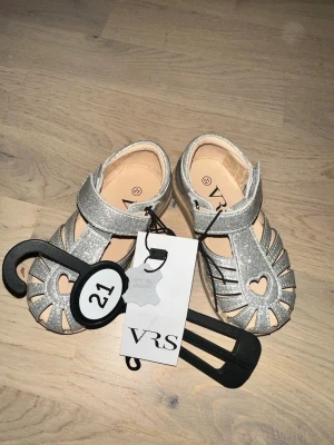 VRS Sandaler, Silver, EUR 21 Glittriga silvriga sandaler - Söta silverfärgade sandaler med hjärtformad detalj. Perfekt för små fötter. Snygga sandaler i silver med glitter från VRS. Skorna har en hjärtformad detalj framtill, kardborreband och öppen tå. Perfekta för sommaren och lätta att ta på. Tillverkade i skinn med mjuk innersula och flexibel yttersula.