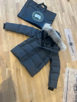 Canada Goose Wyndham Black Label  - Säljer en Canada Goose Wyndham Black Label med fluffig päls på huvan och klassisk patch på ärmen. Jackan har långa ärmar, dragkedja och knappar framtill samt quiltad design. Perfekt för kalla vinterdagar och riktigt snygg streetstil.