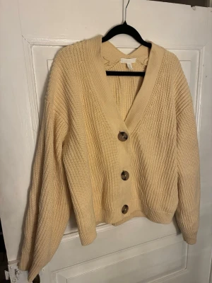 Beige/gul stickad kofta från H&M - Mysig beige/gul stickad kofta från H&M med stora bruna knappar framtill. Koftan har djup v-ringning, breda ribbade muddar och lång ärm. Perfekt för lager-på-lager och chill höst- och vinterstil.