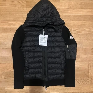 Svart cardigan med huva från Moncler - Svart pufferjacka från Moncler med stickade ärmar och huva. Jackan har quiltad front, dragkedja, ficka med dragkedja på ärmen och Moncler-logga på vänster ärm. Materialmix av polyester och stickad ull ger en snygg och sportig look. Det är en M men kan passa för L