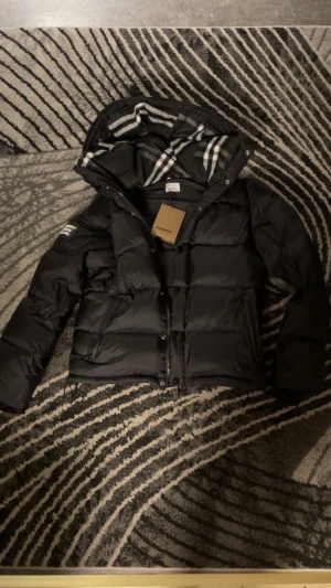 Svart pufferjacka från Burberry - Svart pufferjacka från Burberry med avtagbara ärmar så den kan användas som väst. Jackan har huva med klassiskt rutigt Burberry-foder, stora fickor med dragkedja och tydlig Burberry London England-patch på ärmen. Perfekt för kalla dagar och riktigt snygg streetstil.