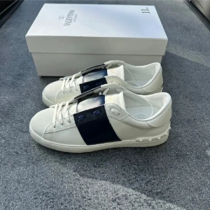 Valentino Garavani open vita sneakers - Snygga vita Valentino Garavani sneakers med bred marinblå panel över sidan och snörning. Skorna har rund tå, platt sula med mönstrad undersida och coola nitar på hälen. Tillverkade i skinn för en lyxig känsla och stilren look. Skorna är använda 4 gånger bara så nästan nya. Pris går att diskutera men inga skambud!