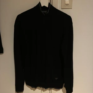 Emporio Armani full zip - Svart kofta från Emporio Armani med dragkedja hela vägen och diskret logga på nedre delen. Långärmad modell med ribbade muddar och grå detaljer vid krage och nederkant. Perfekt för dig som gillar stilrena och klassiska plagg.