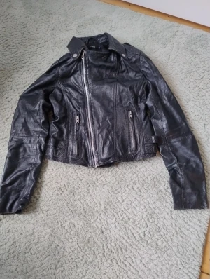 Svart skinnjacka från  - Snygg svart skinnjacka. Jackan har coola quiltade detaljer på ärmarna, dubbla spännen bak och dragkedjor både fram och vid fickorna. Klassisk krage och figurnära passform som ger en edgy vibe.