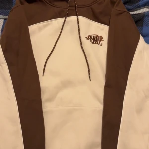 Syna World - Snygg hoodie Syna World i beige och brunt med coolt broderat tryck på bröstet. Tröjan har huva med matchande snören och breda färgblock längs ärmar och axlar. Perfekt för dig som gillar streetwear och vill ha en chill look. Storlek S men passar som M också, priset kan diskuteras lite 