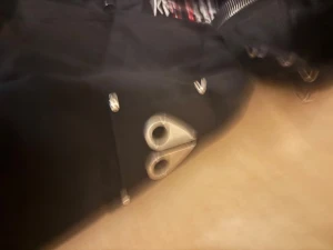 Svart pufferjacka med pälsluva - Säljer en svart pufferjacka från Moose Knuckles med päls på luvan och coola metalldetaljer. Jackan har dragkedja, knappar och två stora silverfärgade öljetter framtill. Insidan har ett snyggt tryck med logga. Perfekt för kalla dagar.