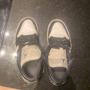 Nike Jordan 1 svart och vit sneakers - Säljer ett par Nike Jordan 1 sneakers i svart och vitt med klassisk snörning och perforerad tå. Skorna har rund tå och platt sula, med den ikoniska Jumpman-loggan på plösen. Perfekt för dig som gillar streetwear och vill ha en stilren look.