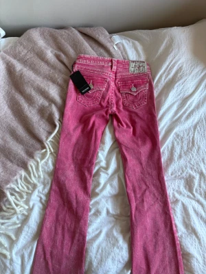 True Religion rosa jeans med kontrastsömmar - Snygga rosa jeans från True Religion med vita kontrastsömmar och ikoniska ficklock bak med silverfärgade knappar. Modellen har klassisk femficksdesign och ett coolt True Religion-märke på linningen. Perfekt för dig som vill sticka ut med färg och detaljer.
